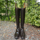 A pair of tall glossy dark brown rain boots standing upright on a gravel path with green foliage in the background.