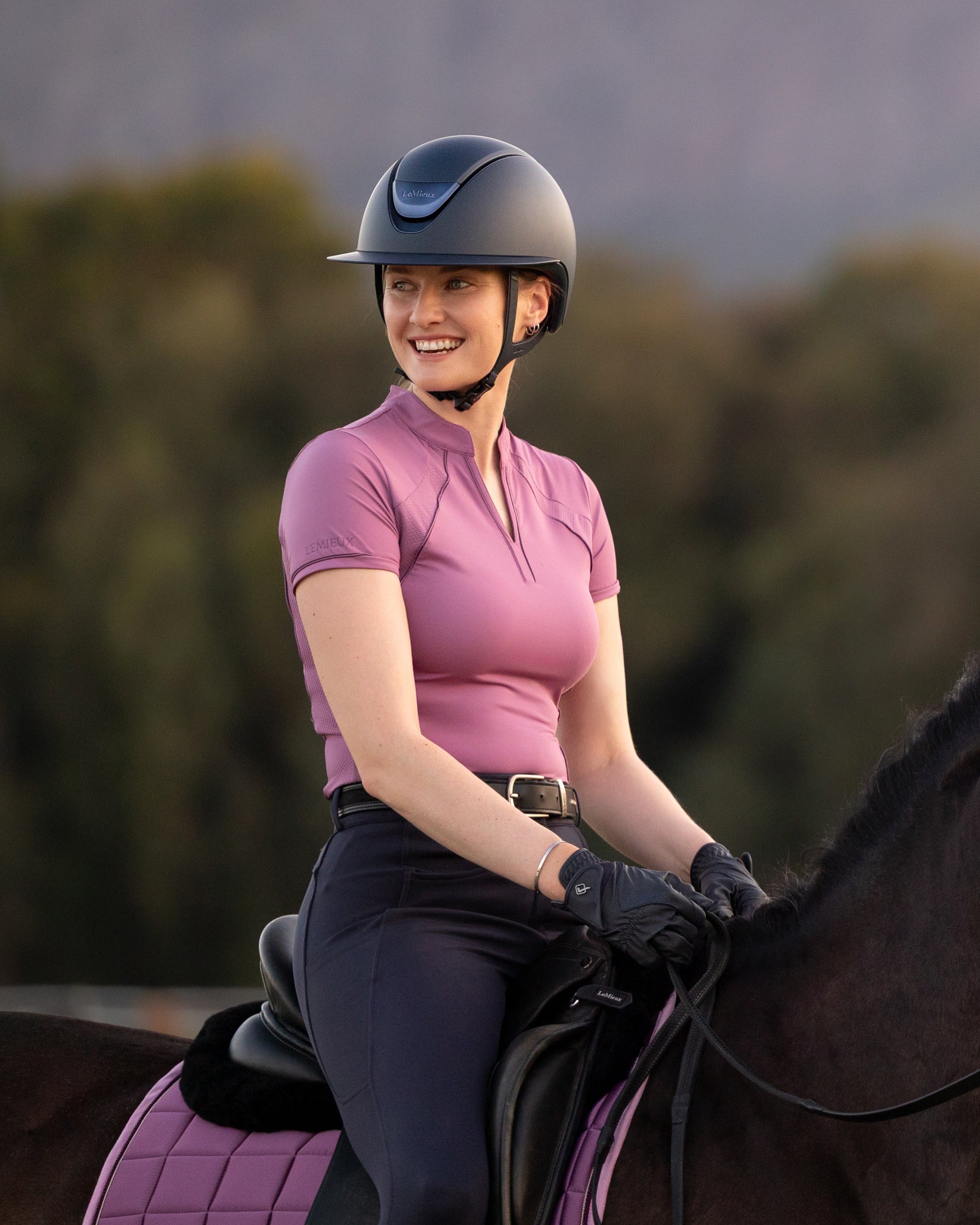 A woman riding a dark brown horse outdoors, wearing a pink riding shirt, black pants, and a dark helmet. The horse is outfitted with a pink fly veil and matching pink saddle pad.