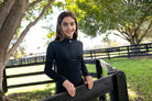 Young girl in a black jacket standing outdoors near a wooden fence with trees and grass in the background.