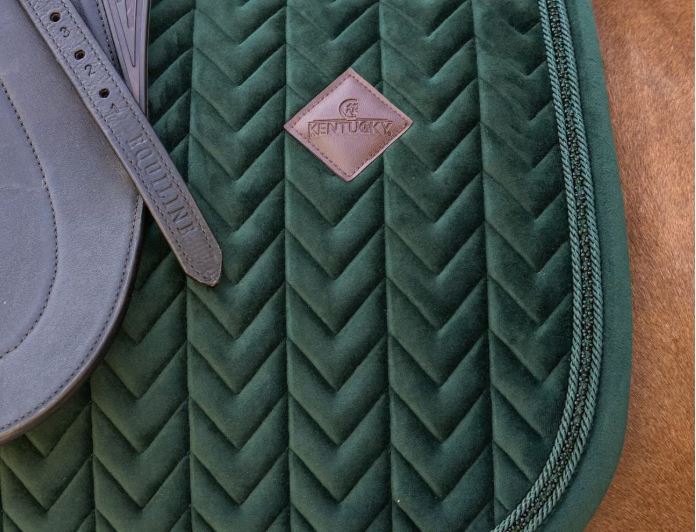 A close-up of a dark green quilted horse saddle pad with a chevron pattern and a brown leather Kentucky brand patch, shown next to a gray saddle on a brown horse blanket.