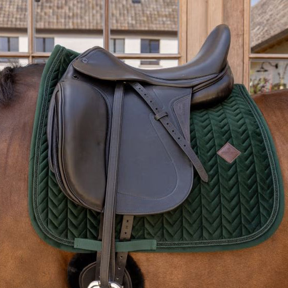A dark green quilted horse saddle pad with a chevron pattern and a small brown diamond logo, shown under a black equestrian saddle on a brown horse.