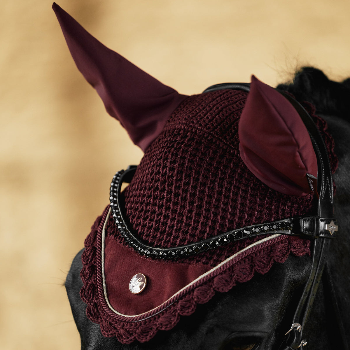 A close-up of a horse wearing a burgundy knit fly veil and matching ear covers with black leather bridle straps against a warm beige background.