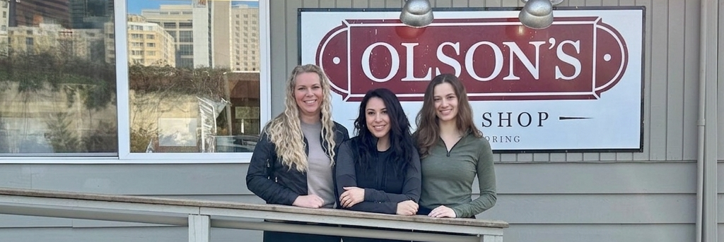Three women standing together on a wooden deck in front of Olson's Shop, wearing casual jackets and pants.