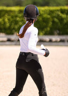 Person in equestrian attire walking on a sandy surface with greenery in the background