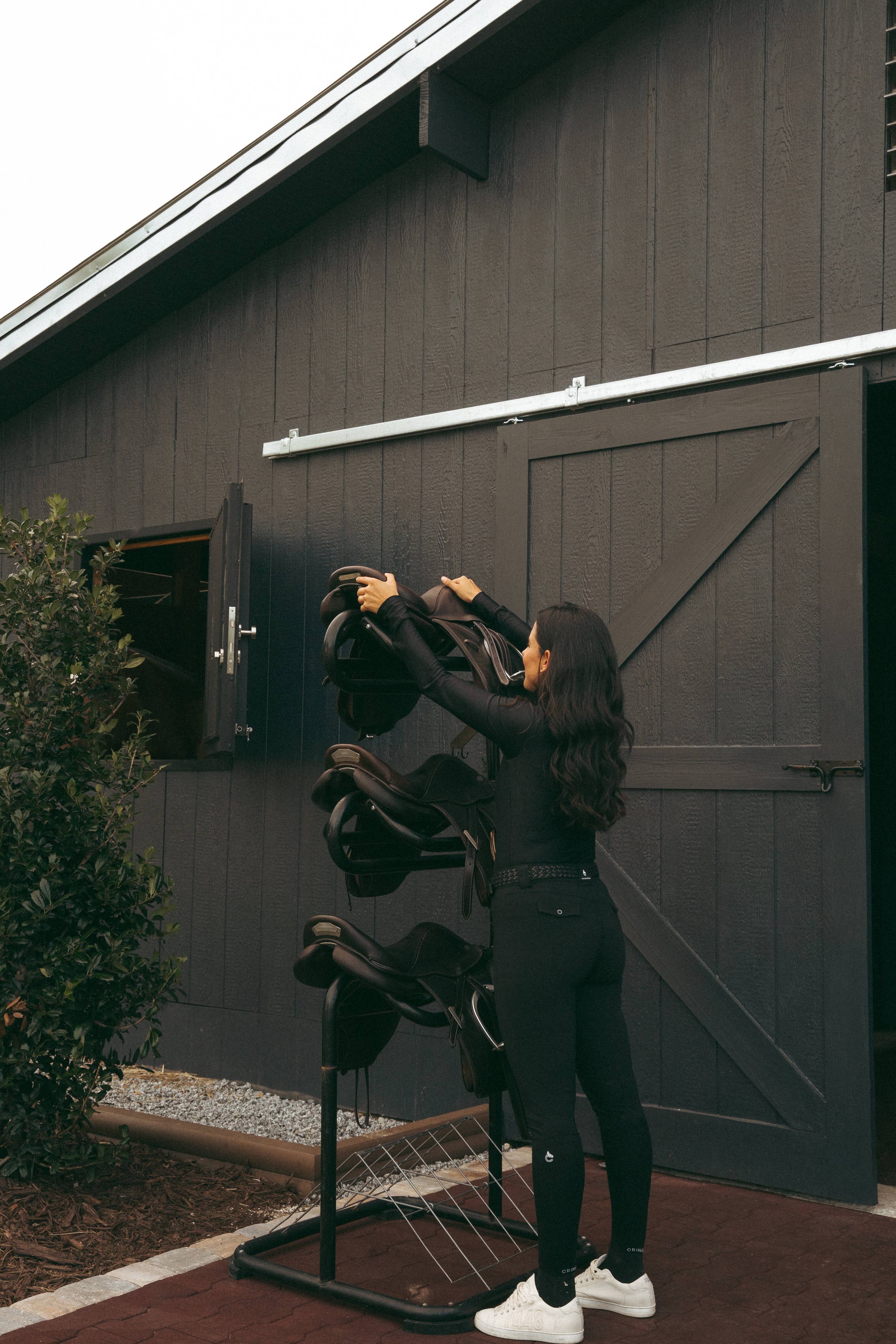 Person organizing horse saddles on a stand in front of a barn.