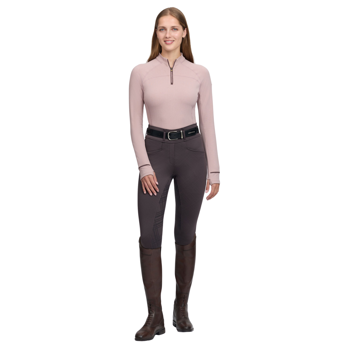 A woman modeling a light pink long-sleeve quarter-zip top, dark gray riding breeches, a black belt, and dark brown riding boots against a plain white background.