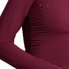 Close-up of a long-sleeve ruched top in deep burgundy, showing the side seam detail and fabric texture against a light gray background.