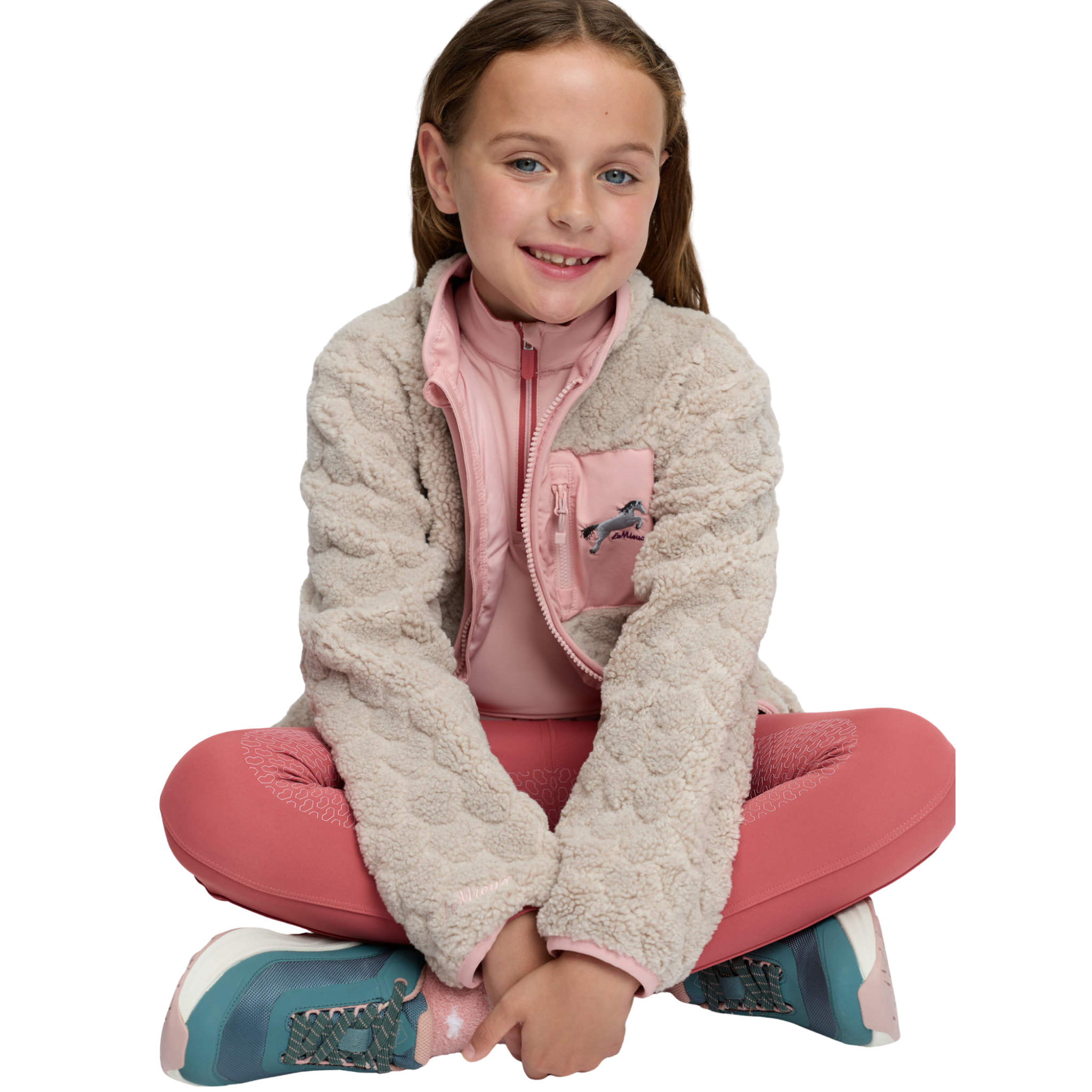 A young girl with light brown hair sits cross-legged against a solid white background, wearing a textured cream fleece zip-up jacket over a pink zip-up top, pink leggings, and teal sneakers.
