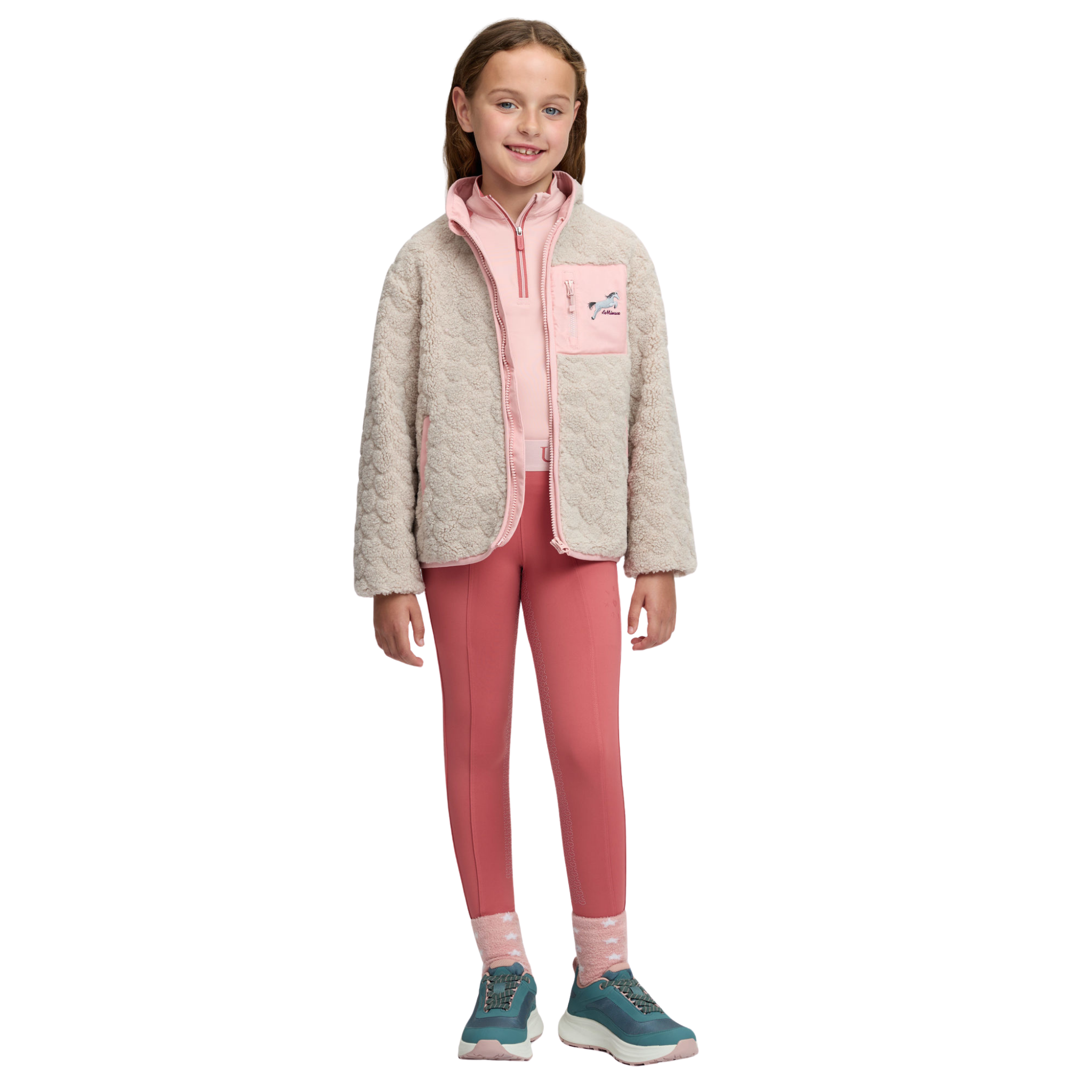 A young girl stands against a white studio background wearing a light beige quilted zip-up jacket with pink pockets over a pink top, pink leggings, pink socks, and teal sneakers.