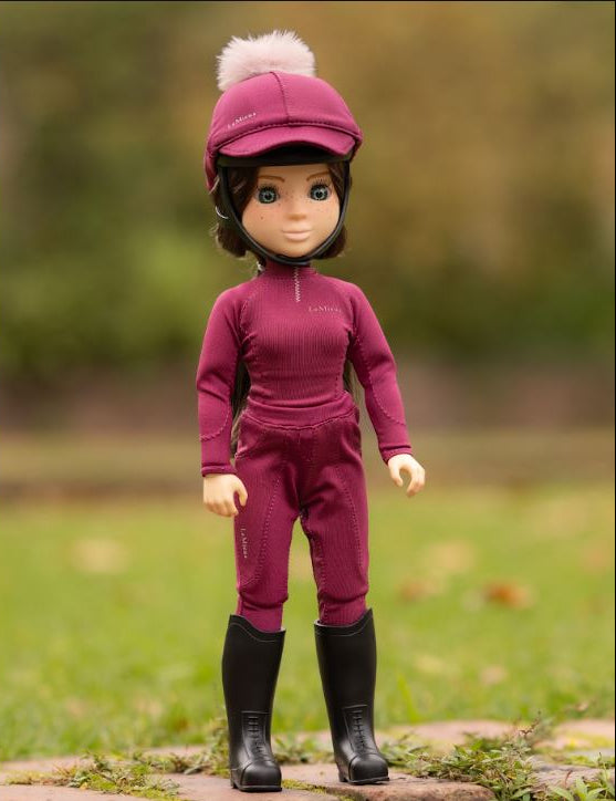 A doll with a brown face and short dark hair stands outdoors on a stone path wearing a matching burgundy riding outfit with a hat and fluffy pom-pom, plus black boots, against a blurred green background.