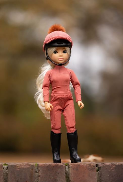 A doll with long white hair wearing a dusty rose riding suit, matching helmet with a brown pom-pom, and black riding boots, standing on a brick ledge with a blurred outdoor background.