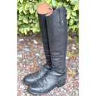 A pair of tall black leather riding boots standing upright on a gravel path with green foliage in the background.