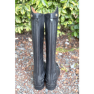 A pair of tall black leather riding boots with side zippers, standing upright on a gravel surface with green foliage in the background.