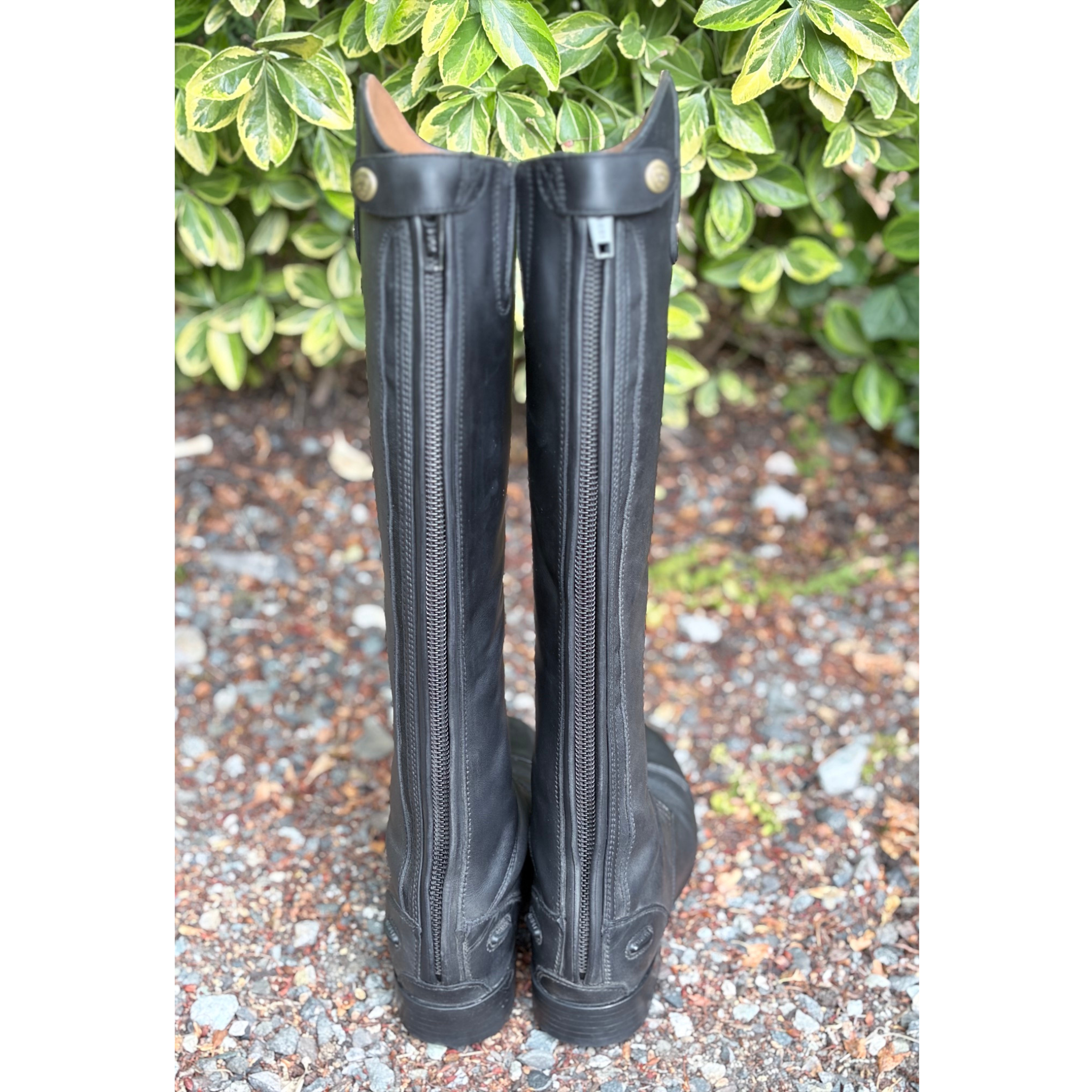 A pair of tall black leather riding boots with side zippers, standing upright on a gravel surface with green foliage in the background.