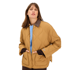 A woman wearing a quilted tan jacket with dark brown collar, cuffs, and pockets, layered over a light blue top, photographed against a solid white background.