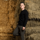 Man in a black sweater and green pants standing among hay bales.
