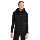 A woman modeling a black hooded zip-front puffer vest over a black top and black pants, photographed against a plain white studio background.
