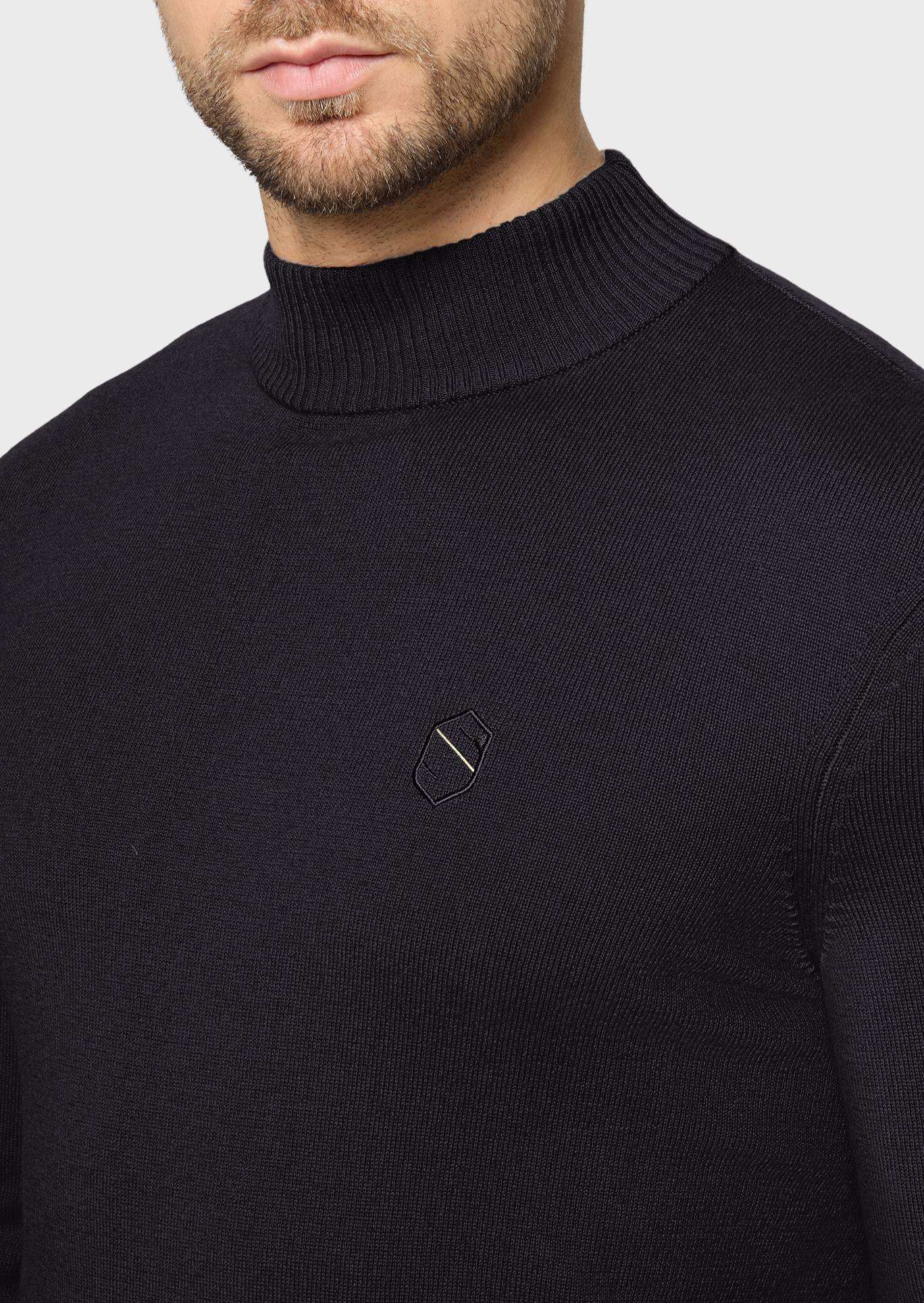 Close-up of a man wearing a black ribbed mock neck sweater with a small geometric embroidered logo on the left chest, against a light gray background.