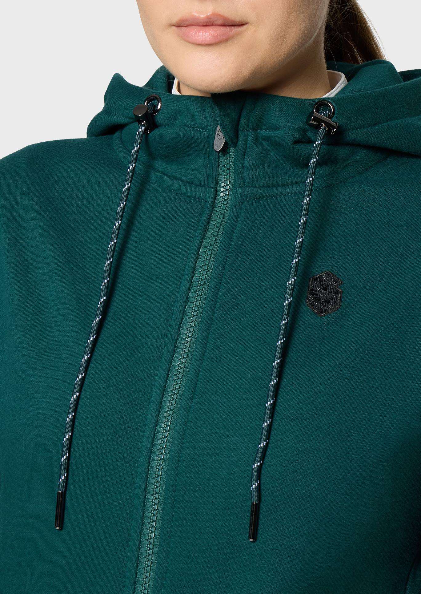 Close-up of a person wearing a dark green zip-up hoodie with black drawstrings and a small embroidered shield logo on the chest.