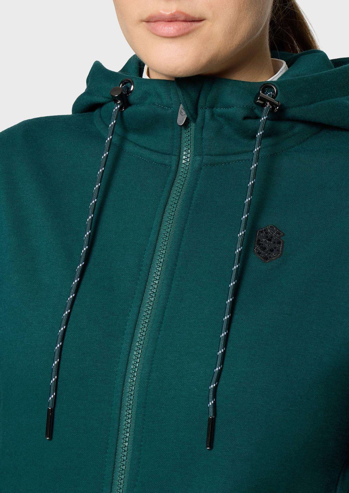 Close-up of a person wearing a dark green zip-up hoodie with black drawstrings and a small embroidered shield logo on the chest.