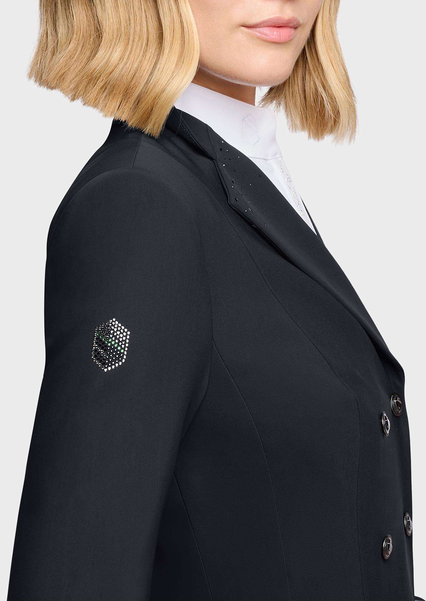 Close-up of a model wearing a black double-breasted blazer with a small rhinestone-embellished crest patch on the left sleeve, paired with a white high-neck shirt.