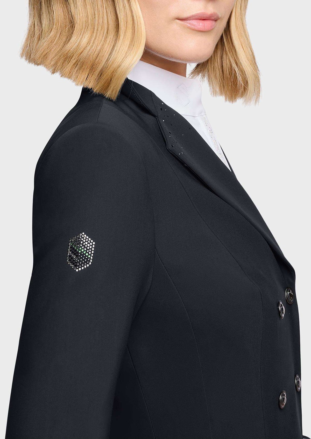 Close-up of a model wearing a black double-breasted blazer with a small rhinestone-embellished crest patch on the left sleeve, paired with a white high-neck shirt.