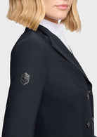 Close-up of a model wearing a black double-breasted blazer with a small rhinestone-embellished crest patch on the left sleeve, paired with a white high-neck shirt.