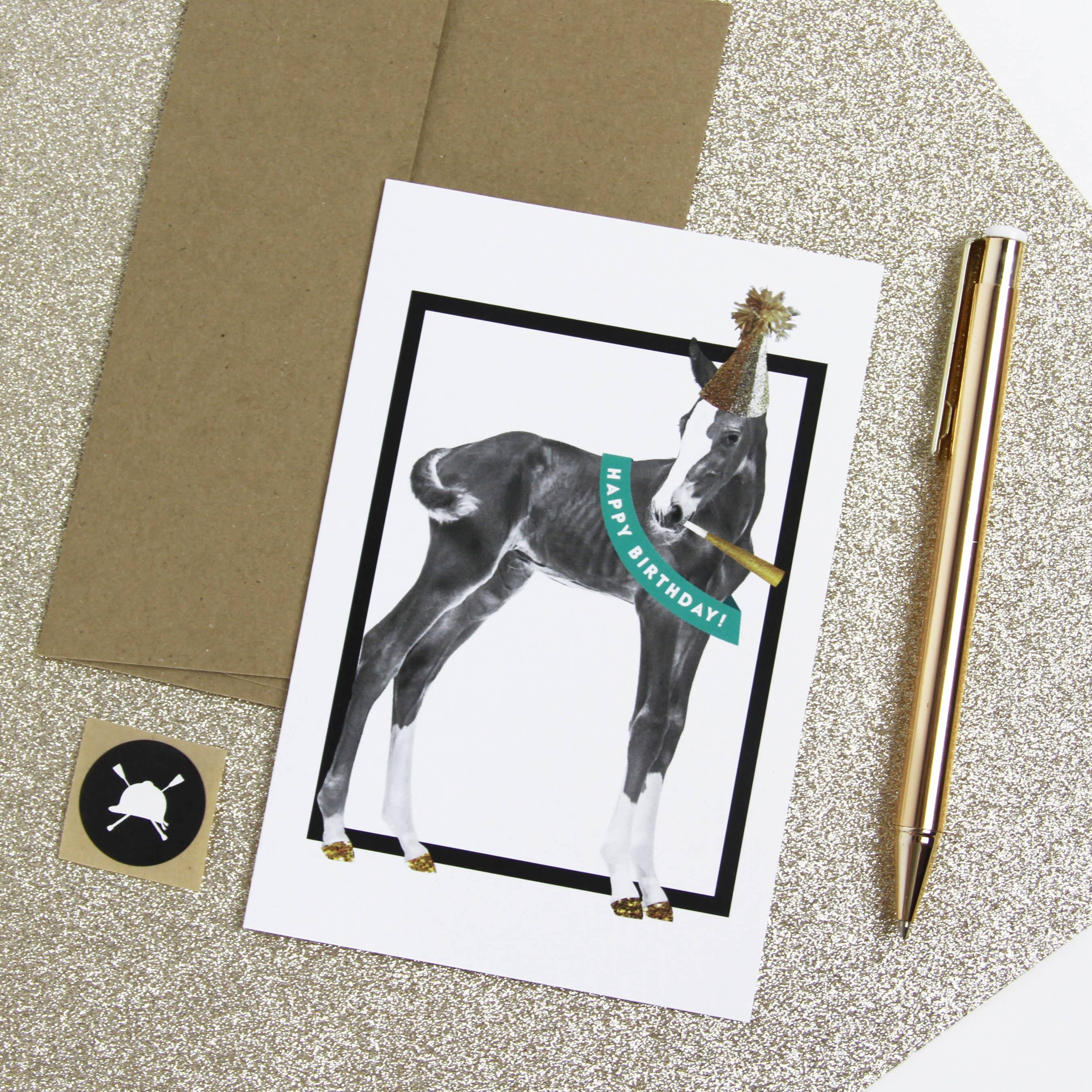 Greeting card with a dog illustration and 'Happy Birthday!' text, placed on a glittery surface with a pen and envelope.