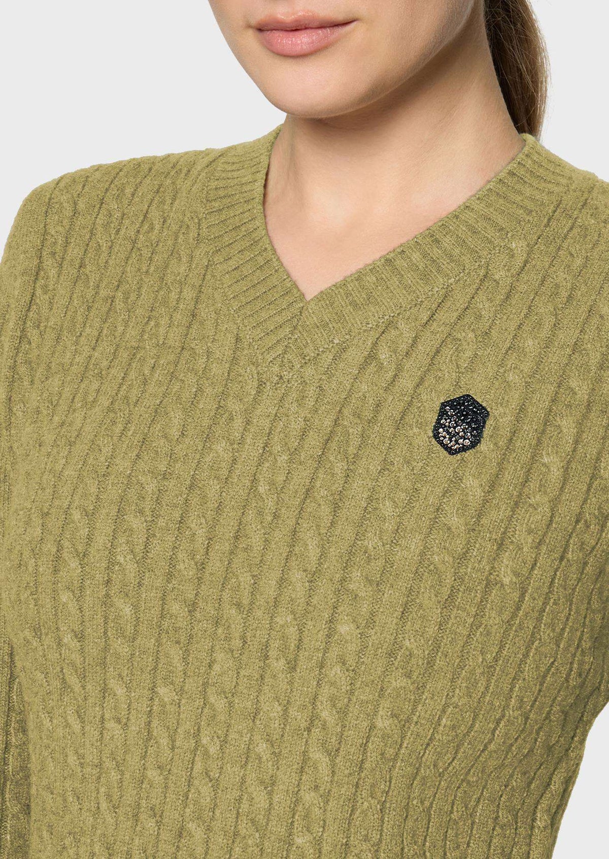 Close-up of a woman wearing a ribbed V-neck sweater in olive green with a small black embroidered shield logo on the left chest.