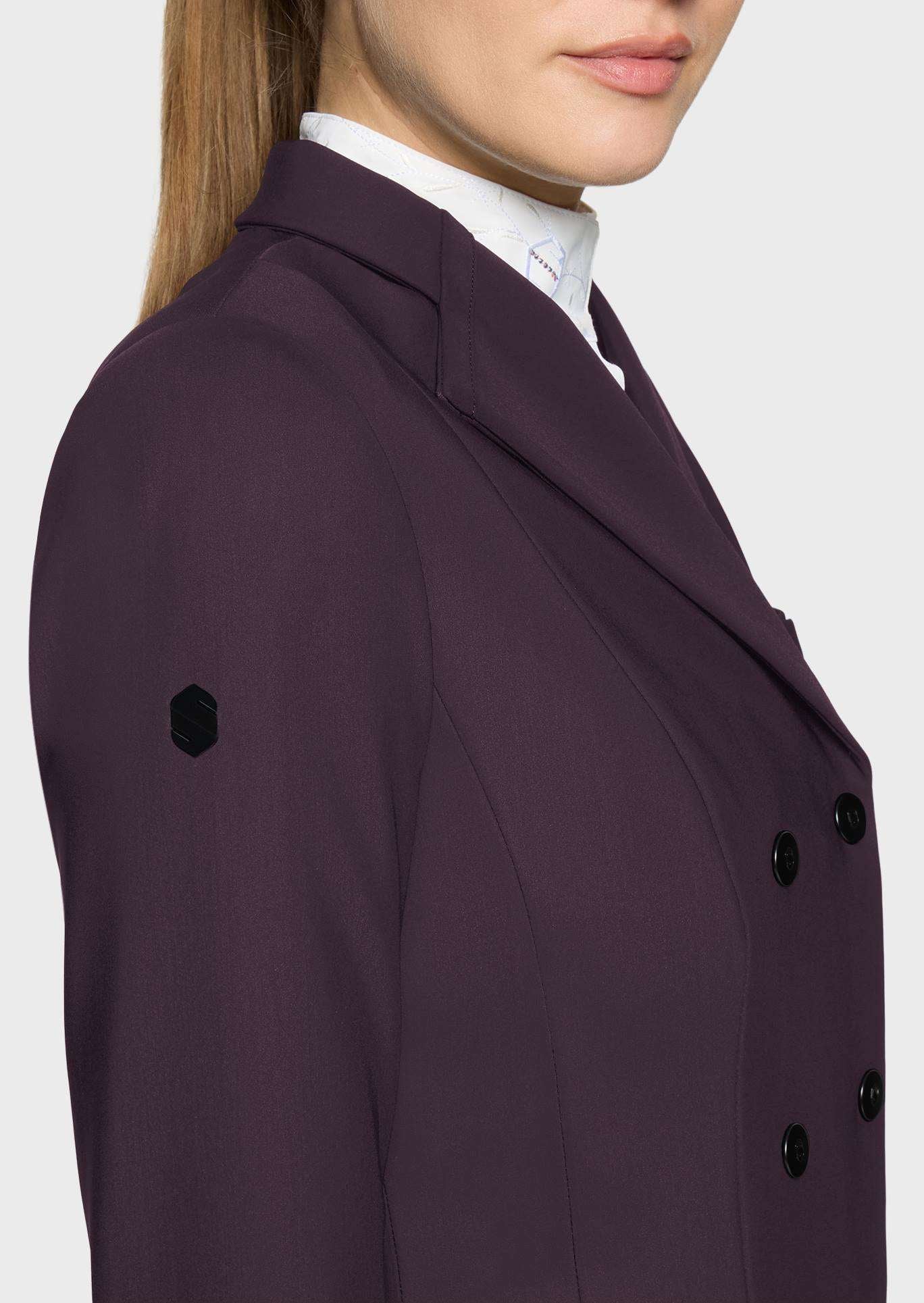 A close-up side view of a model wearing a dark plum purple double-breasted blazer over a white collared shirt, set against a plain light gray background.