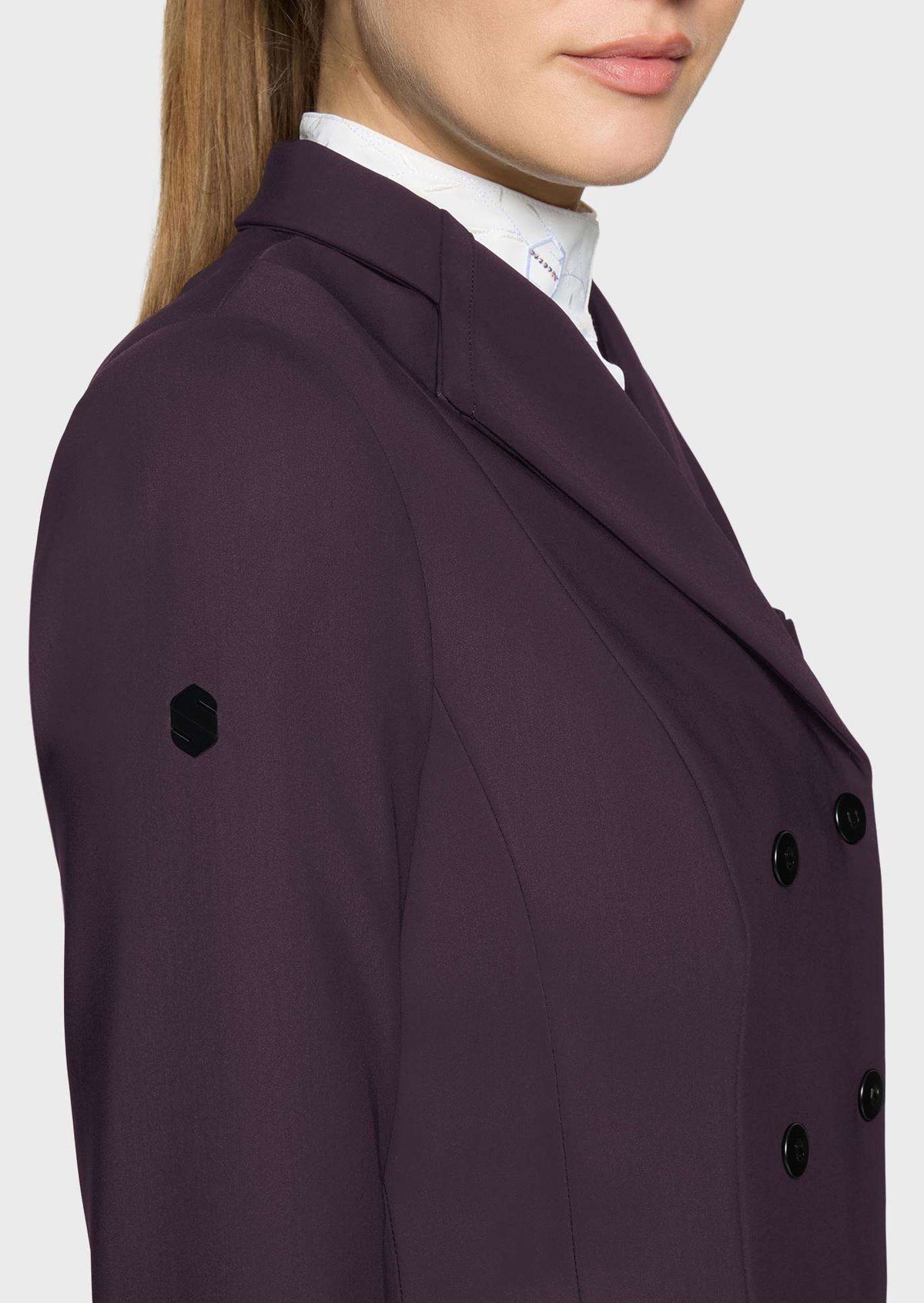 A close-up side view of a model wearing a dark plum purple double-breasted blazer over a white collared shirt, set against a plain light gray background.