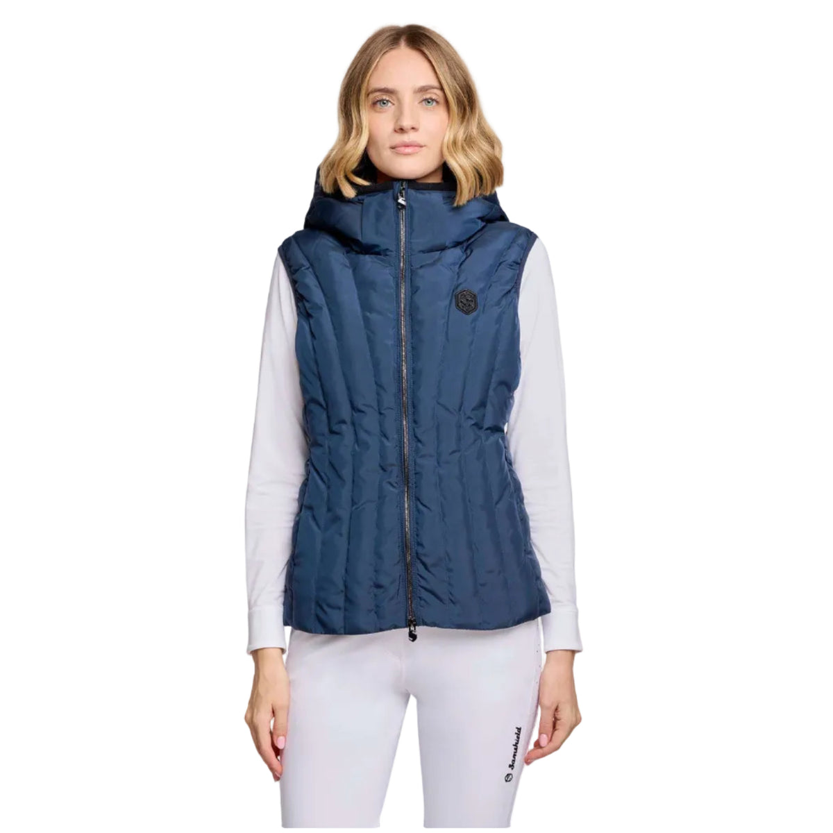 A woman modeling a sleeveless navy blue puffer vest with a front zipper and hood, worn over a white long-sleeve top and white leggings against a light gray studio background.