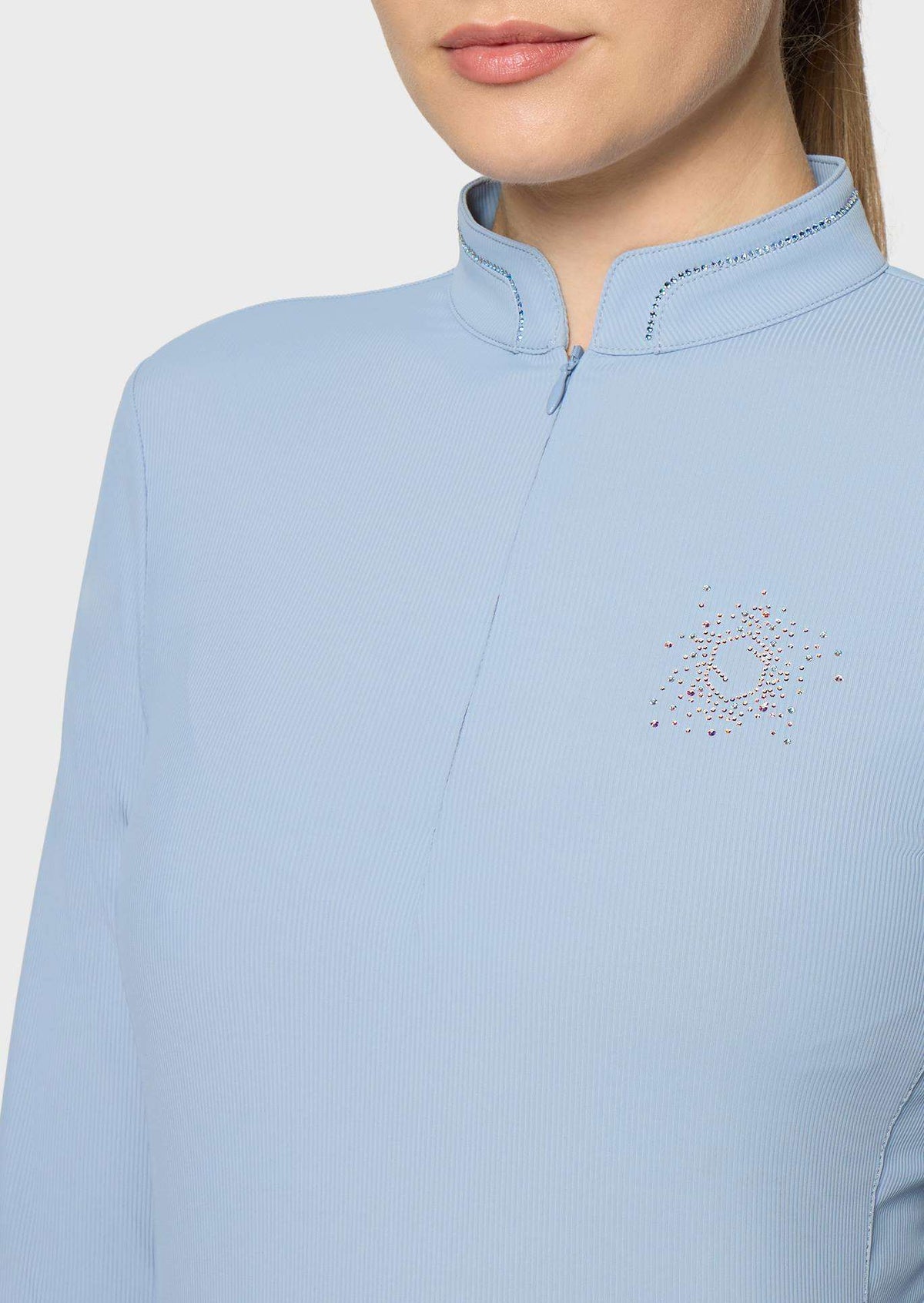 A close-up of a model wearing a light blue ribbed long-sleeve top with a mock neck and a small multicolor rhinestone graphic on the left chest, set against a plain light gray background.