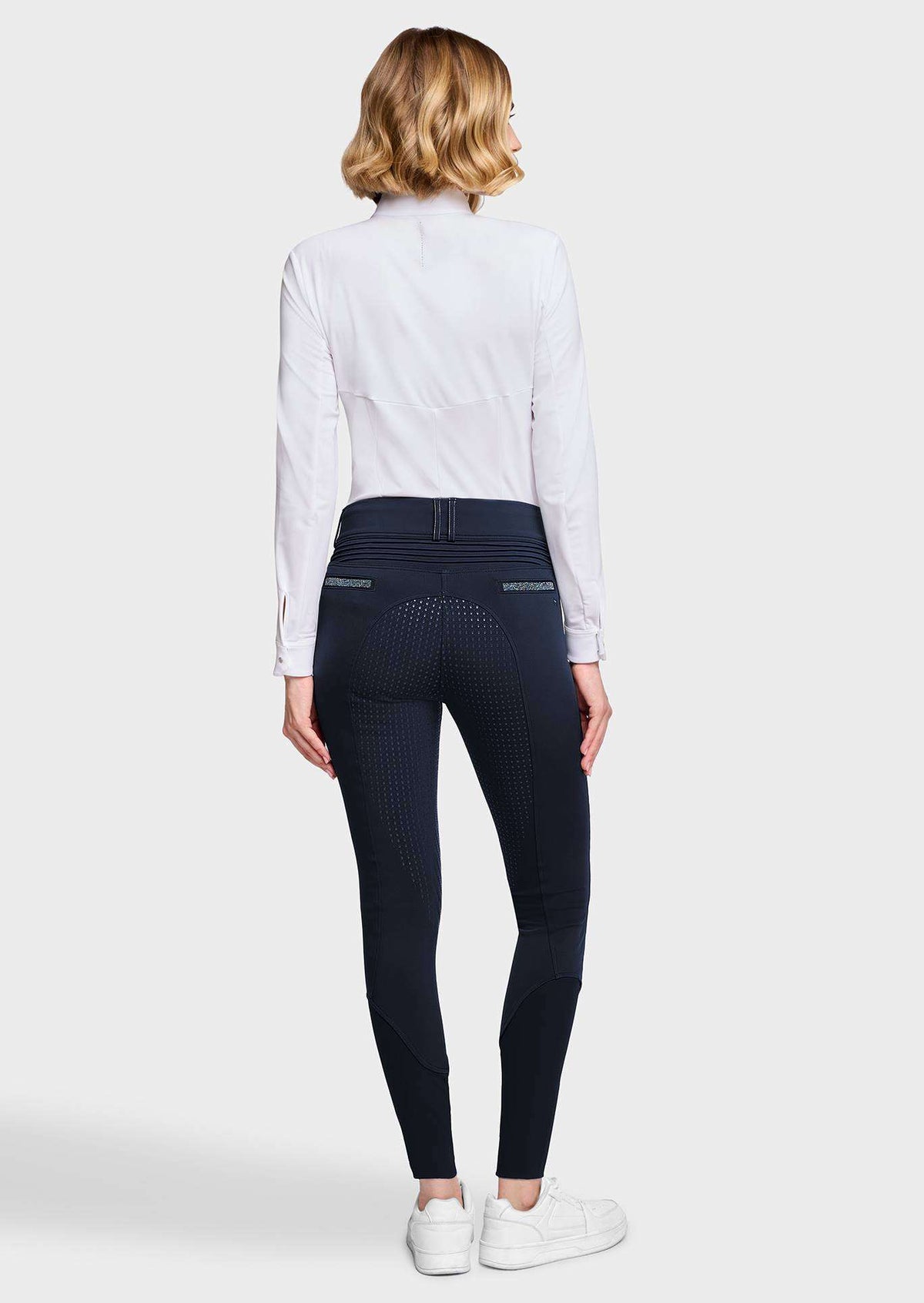 A woman shown from the back wearing a white long-sleeve shirt, dark navy textured riding breeches, and white sneakers against a light gray studio background.