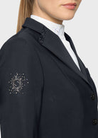 Close-up of a woman wearing a dark navy tailored blazer with decorative crystal embellishments on the shoulder and sleeve, over a white collared shirt, against a light gray studio background.