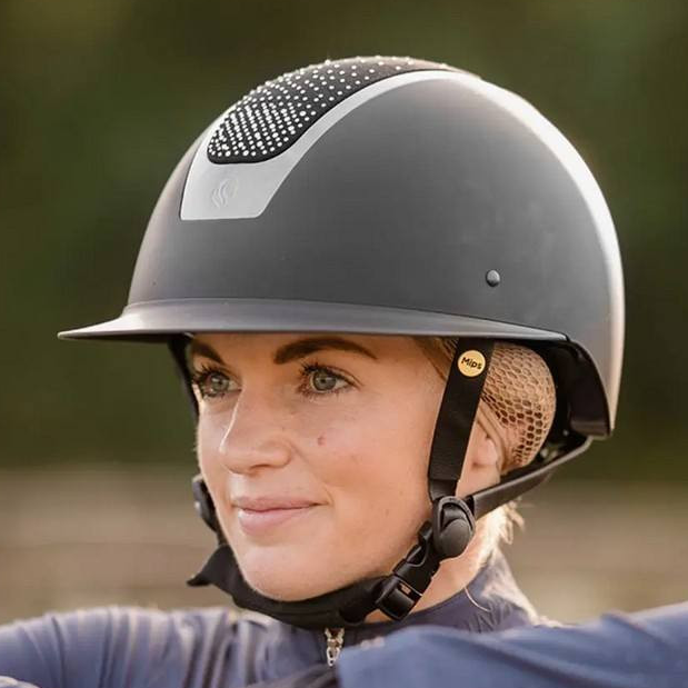 Person wearing a gray riding helmet and gloves, leaning on a wooden fence with a blurred natural background
