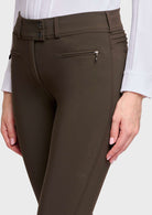Close-up of a person wearing dark brown equestrian riding tights with a zippered side pocket and a subtle embossed logo on the thigh, paired with a white shirt.