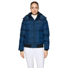 A woman modeling a navy blue puffer jacket with a black zipper, black trim at the hem and cuffs, and a small embroidered crest on the chest, paired with white pants against a light gray studio background.