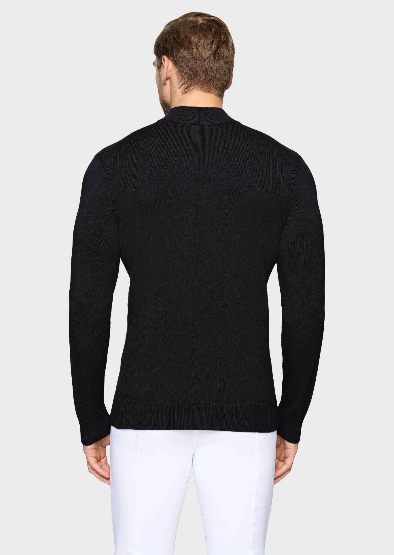 A man viewed from the back wearing a black long-sleeve knit sweater and white trousers, standing against a light gray studio background.