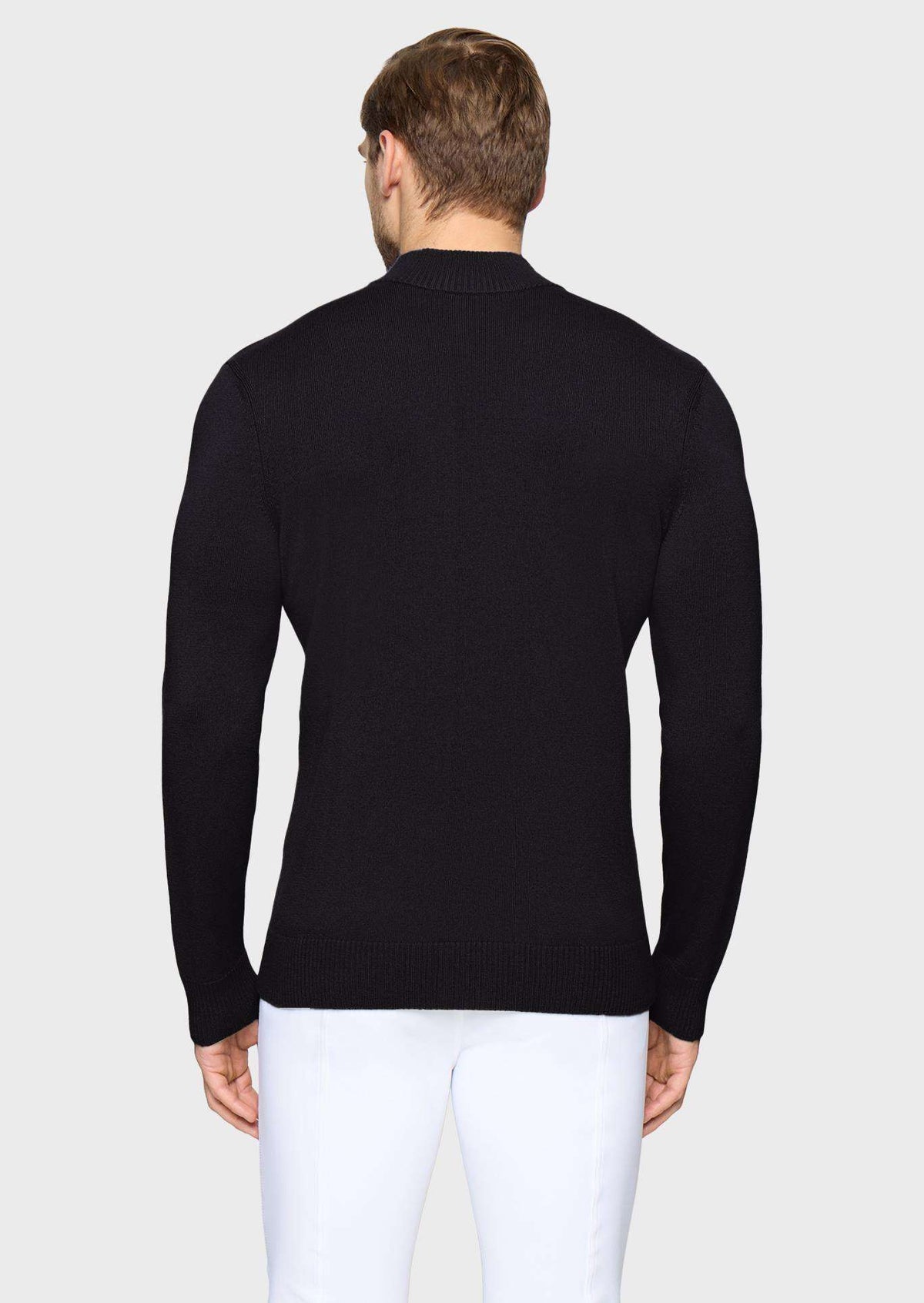A man viewed from the back wearing a black long-sleeve knit sweater and white trousers, standing against a light gray studio background.