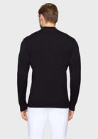 A man viewed from the back wearing a black long-sleeve knit sweater and white trousers, standing against a light gray studio background.
