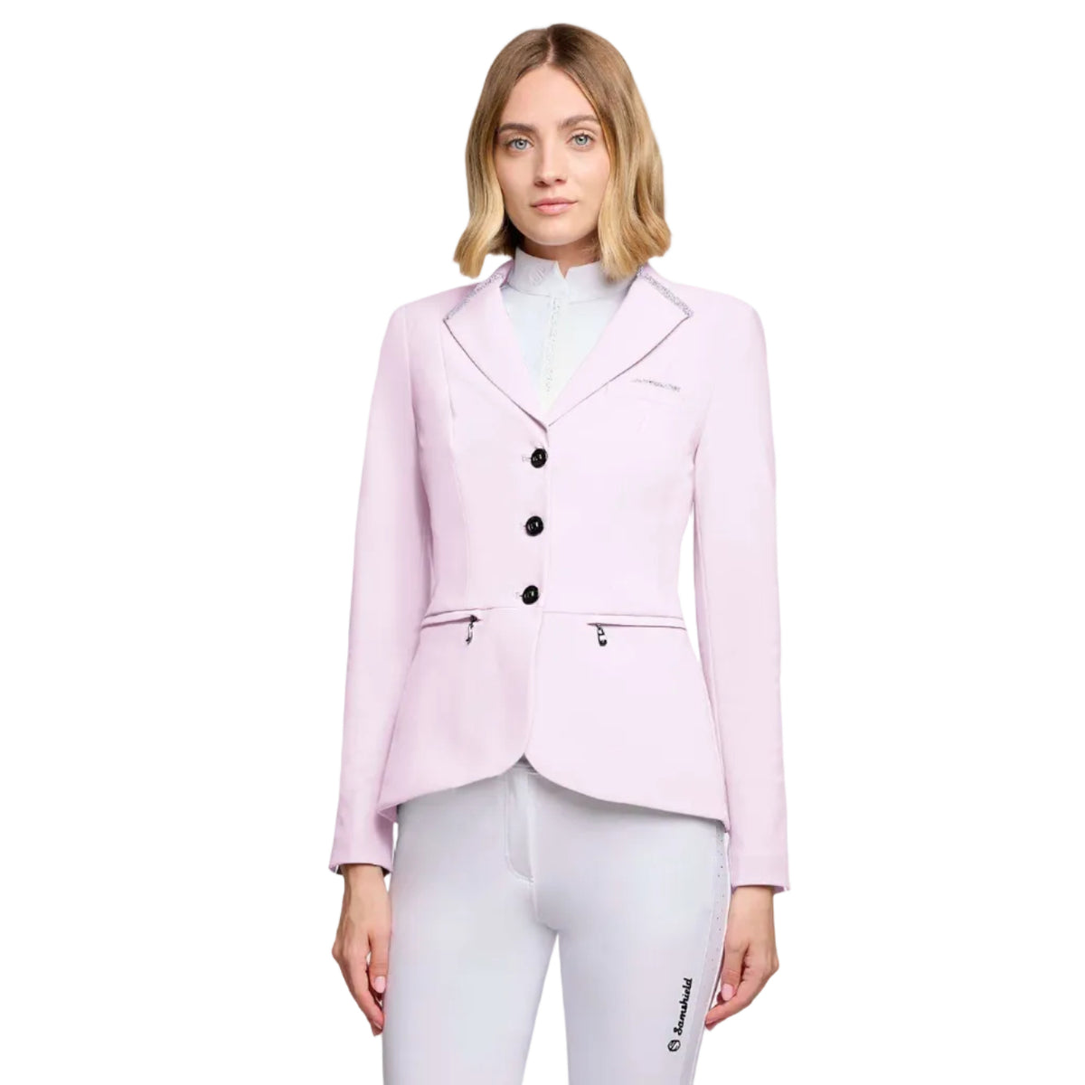 A woman modeling a light pink equestrian-style blazer with black buttons and silver piping, paired with white riding pants, against a light gray studio background.