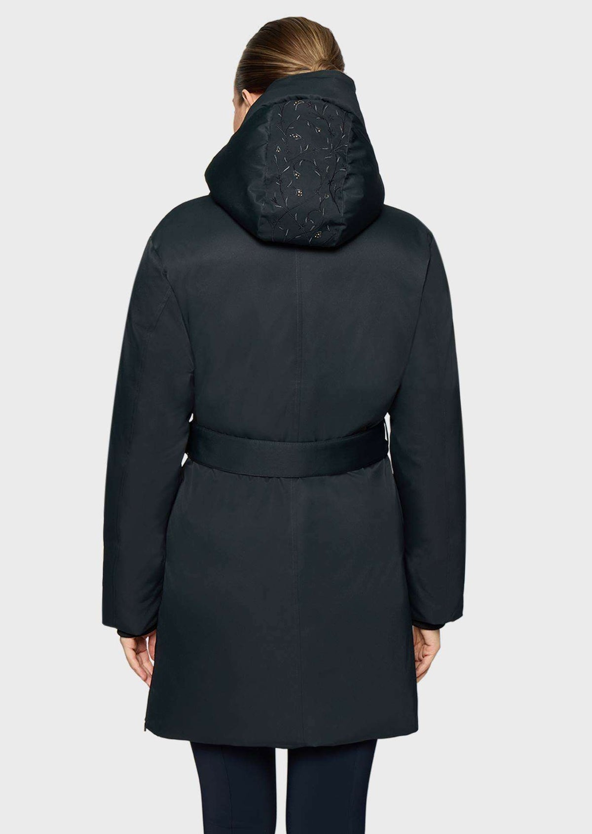 A woman viewed from the back wearing a dark navy hooded puffer coat with a matching waist belt, set against a light gray studio background.