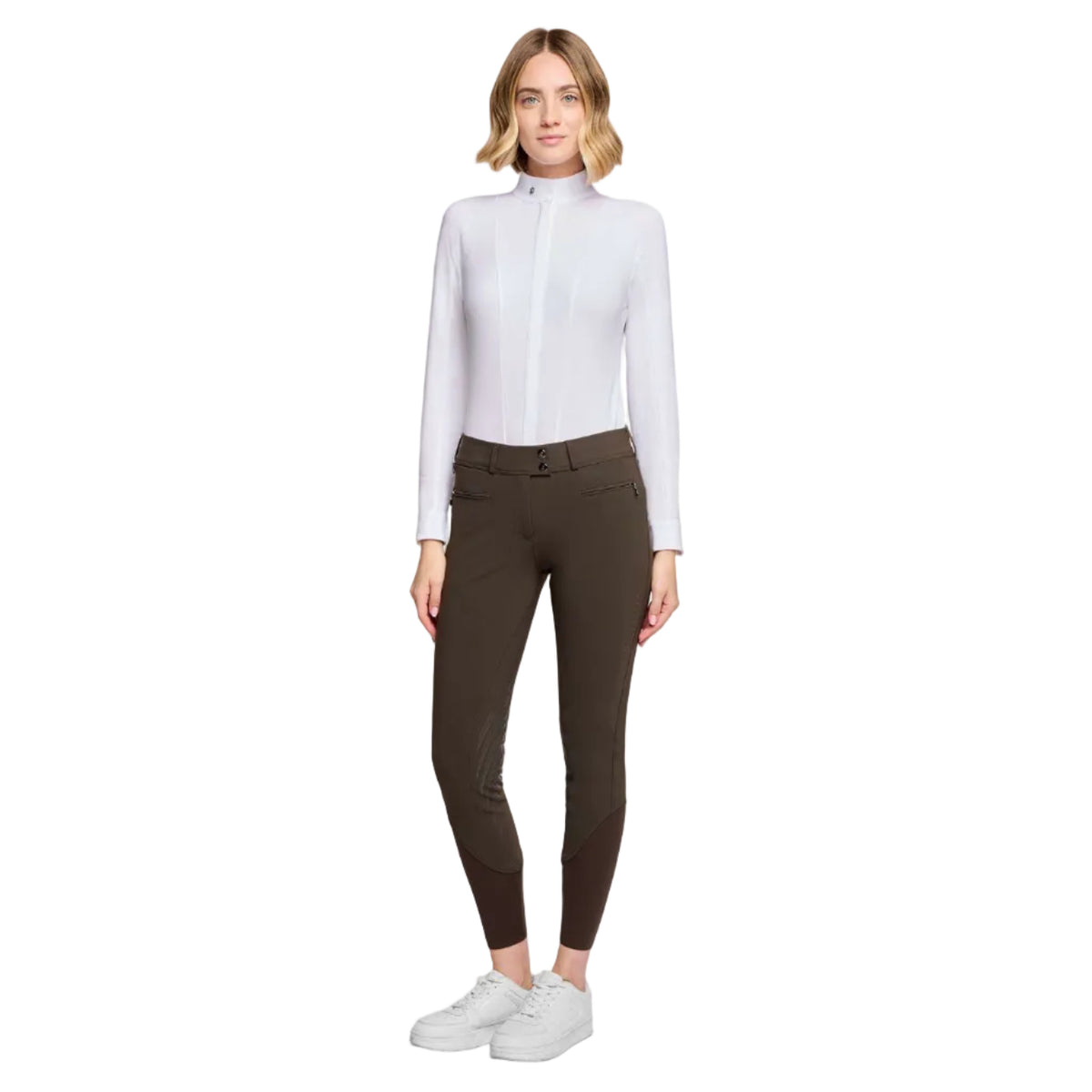 A woman modeling a white long-sleeve mock-neck shirt and dark brown riding breeches with white sneakers, shot against a light gray studio background.