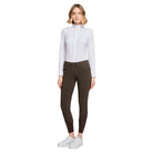 A woman modeling a white long-sleeve mock-neck shirt and dark brown riding breeches with white sneakers, shot against a light gray studio background.