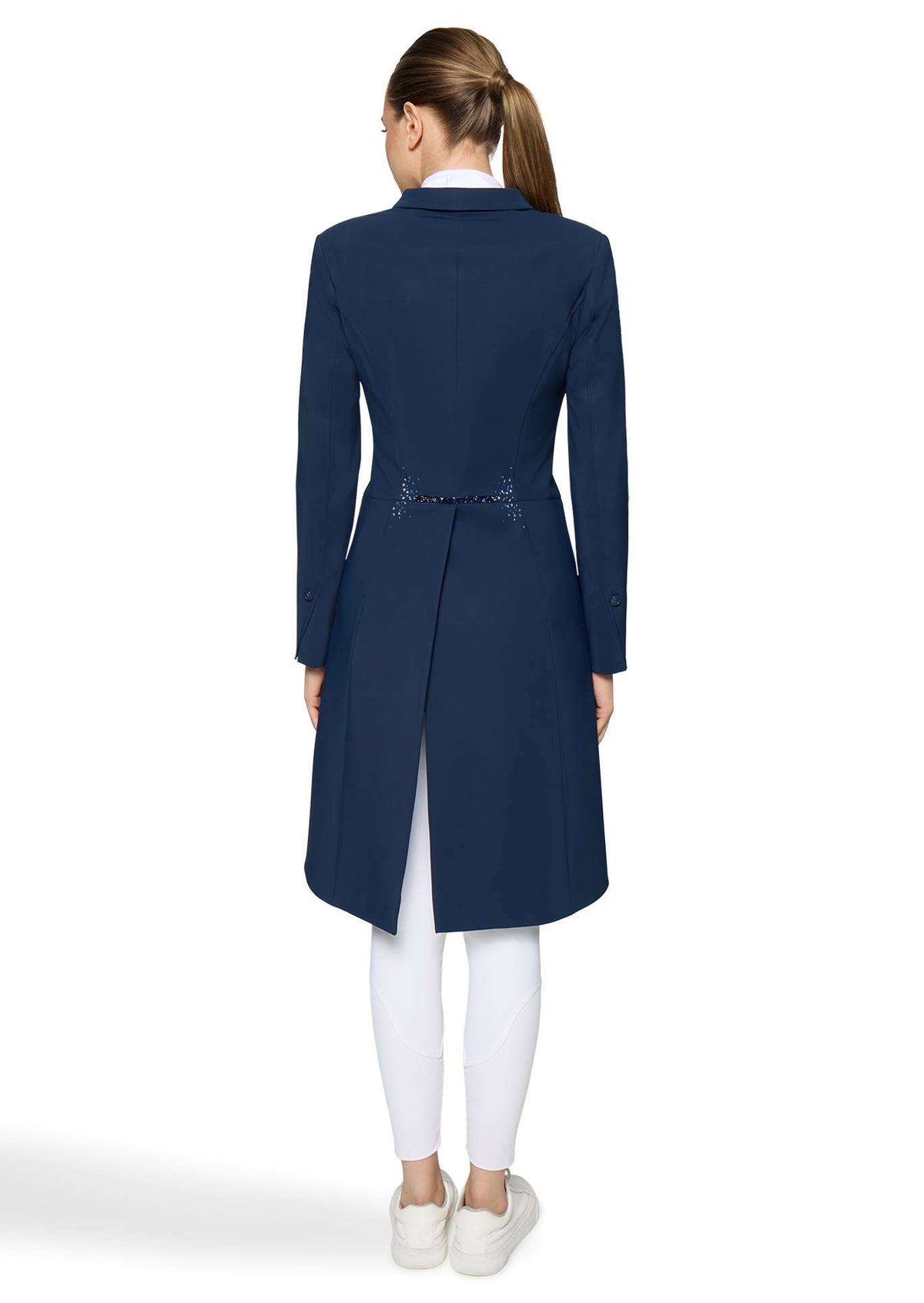 A woman viewed from the back wearing a navy blue long coat over a white collared shirt, white leggings, and white sneakers on a light gray studio background.