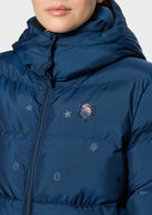 Close-up of a person wearing a navy blue hooded puffer jacket with a front zipper, quilted stitching, and small embroidered logo details on a light gray background.