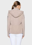 A woman viewed from the back wearing a light taupe hooded jacket and white pants against a plain light gray studio background.
