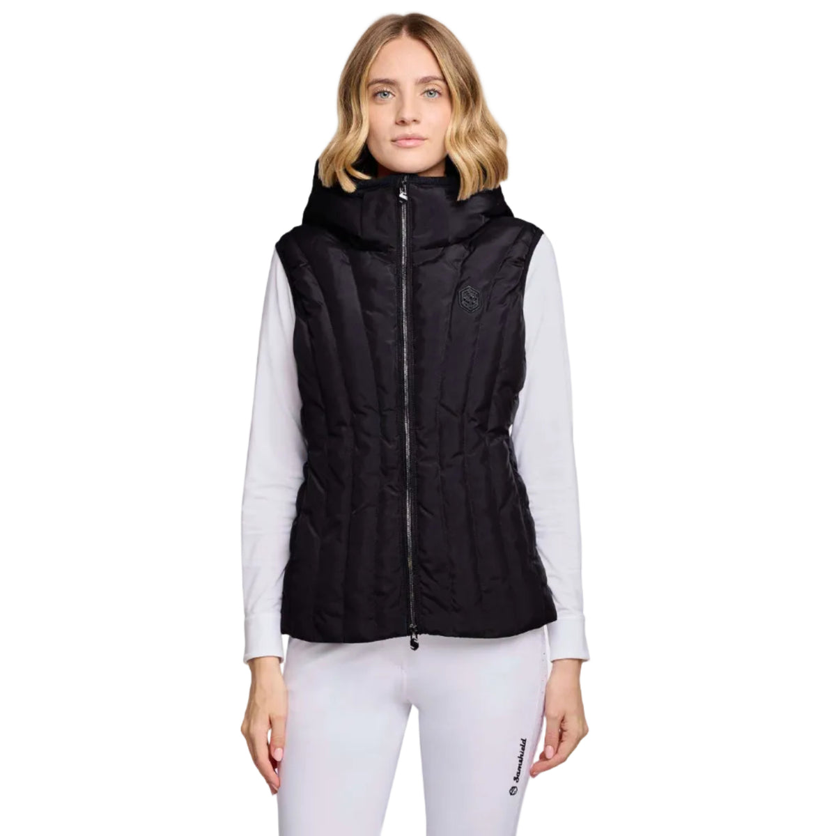 A woman modeling a black sleeveless puffer vest with a high collar and front zipper, worn over a white long-sleeve top and white leggings, against a light gray studio background.