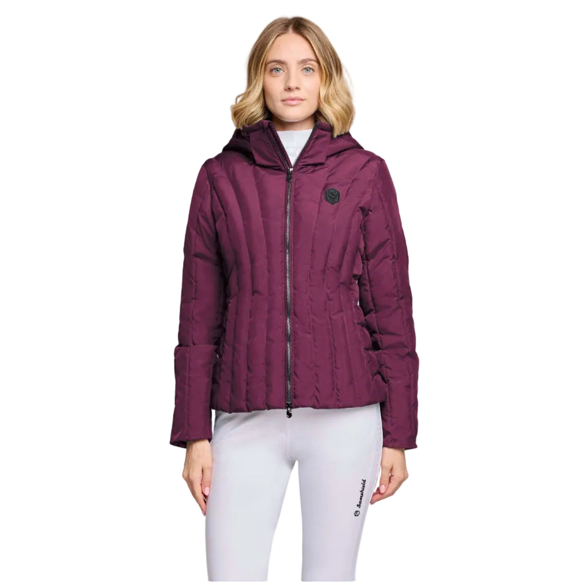 A woman modeling a quilted puffer jacket in deep burgundy with a black zipper and a small circular chest logo, paired with white leggings, against a light gray studio background.